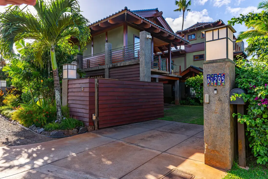 Una casa de varios niveles está detrás de una entrada para vehículos, rodeada de plantas tropicales.