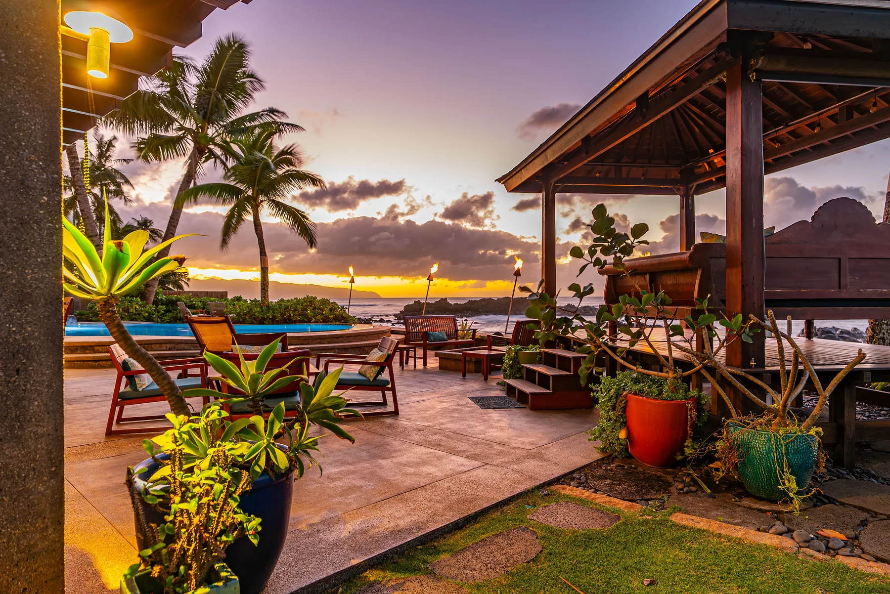 Un patio tropical con asientos de descanso y una pérgola cubierta mira al agua al atardecer.