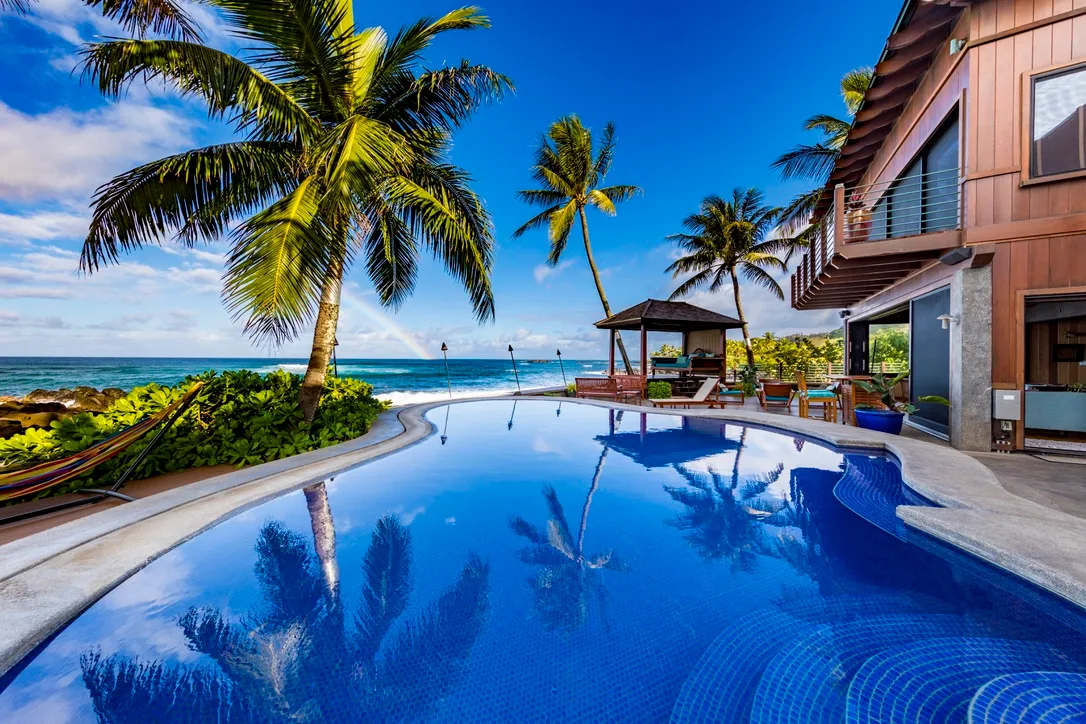 Una zona de piscina tropical junto a la playa, con palmeras y un pabellón.