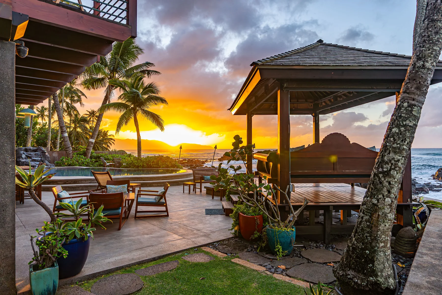 Un patio al aire libre con comedor y asientos cubiertos ofrece vistas al océano al atardecer.