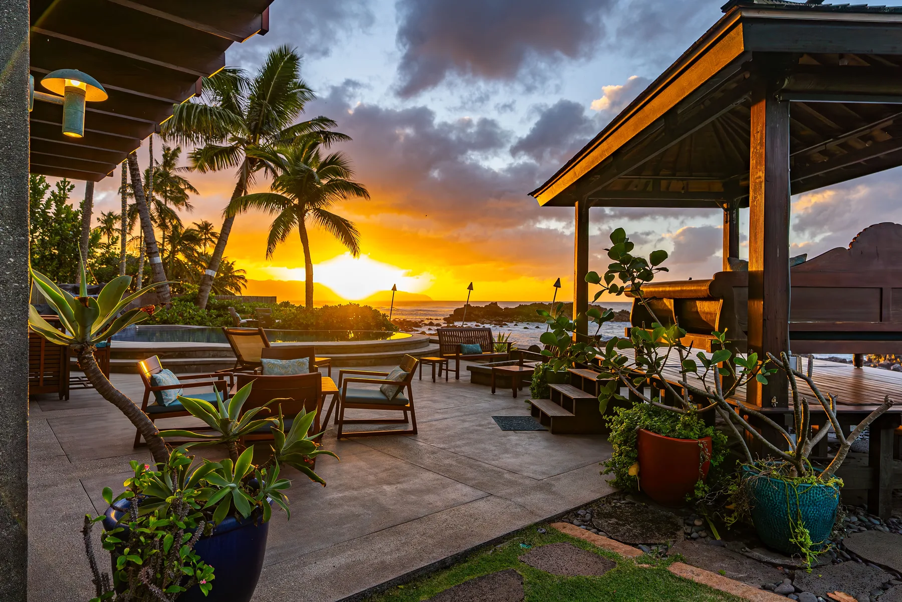 Un patio tropical al aire libre con asientos y plantas en macetas mira hacia el agua al atardecer.