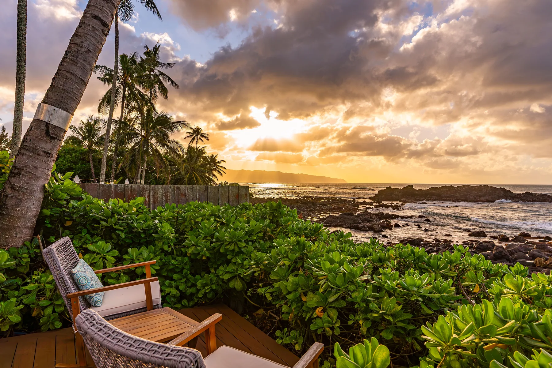 Una costa tropical al atardecer, con palmeras, vegetación frondosa y orilla rocosa.