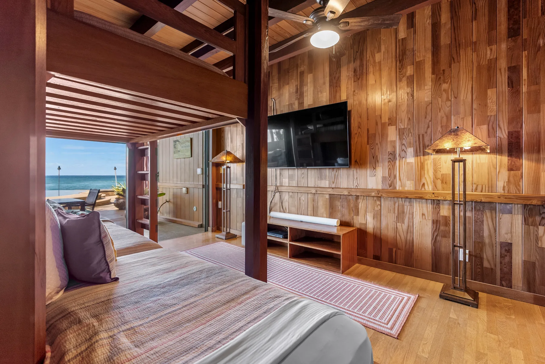 Dormitorio revestido de madera con una cama, un televisor en la pared y vista al agua.