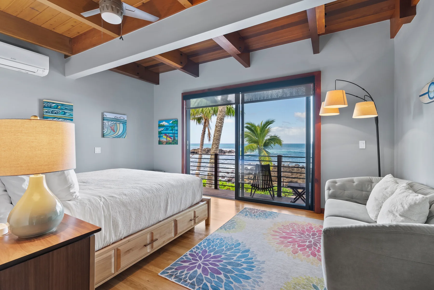 Un dormitorio con cama, asientos y puertas corredizas de vidrio con vista al mar y palmeras.