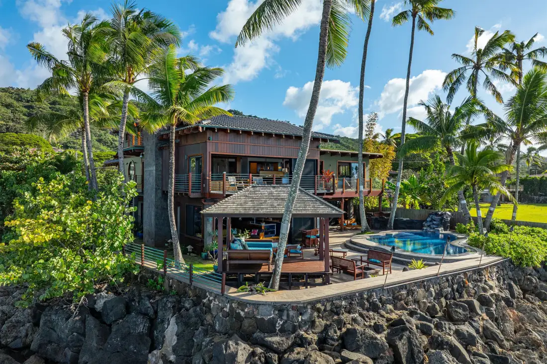Una casa tropical con piscina, terraza y palmeras junto a una costa rocosa.