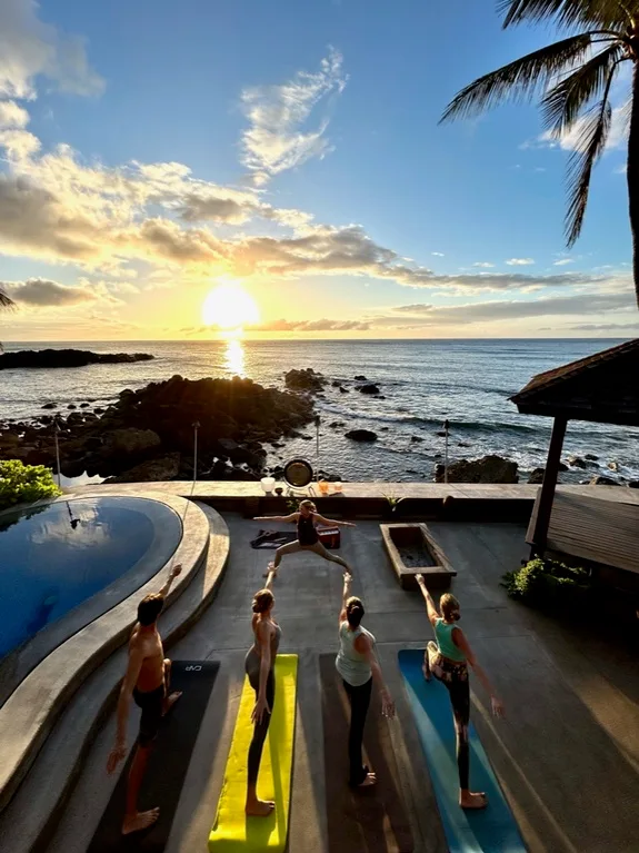 Varias personas practican yoga junto a una piscina con vistas a una costa rocosa al atardecer.