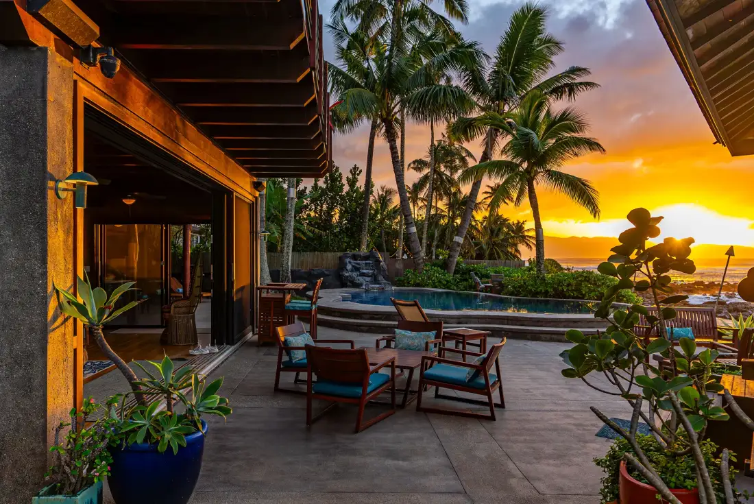 Un patio junto a la piscina con asientos al aire libre mira hacia palmeras y un atardecer.