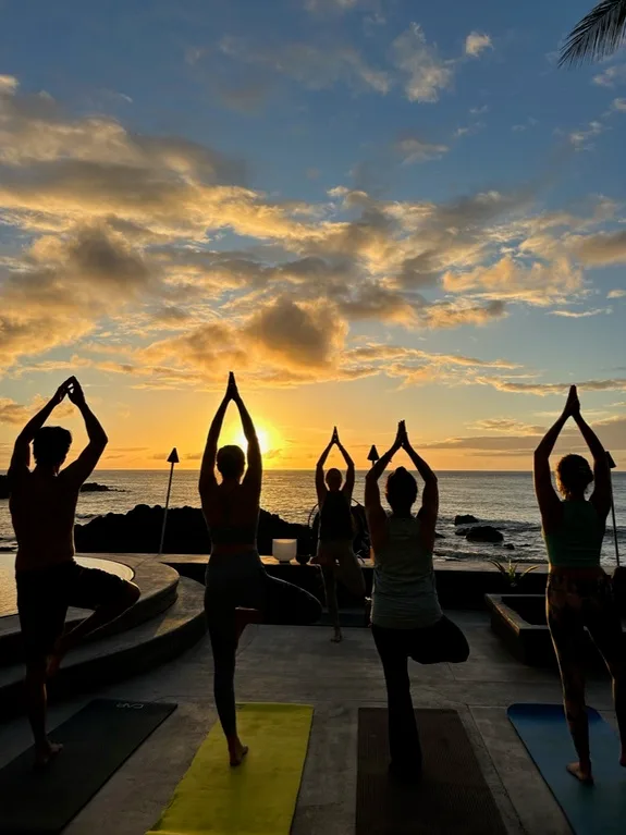 Varias personas practican yoga sobre esterillas junto al mar al atardecer.