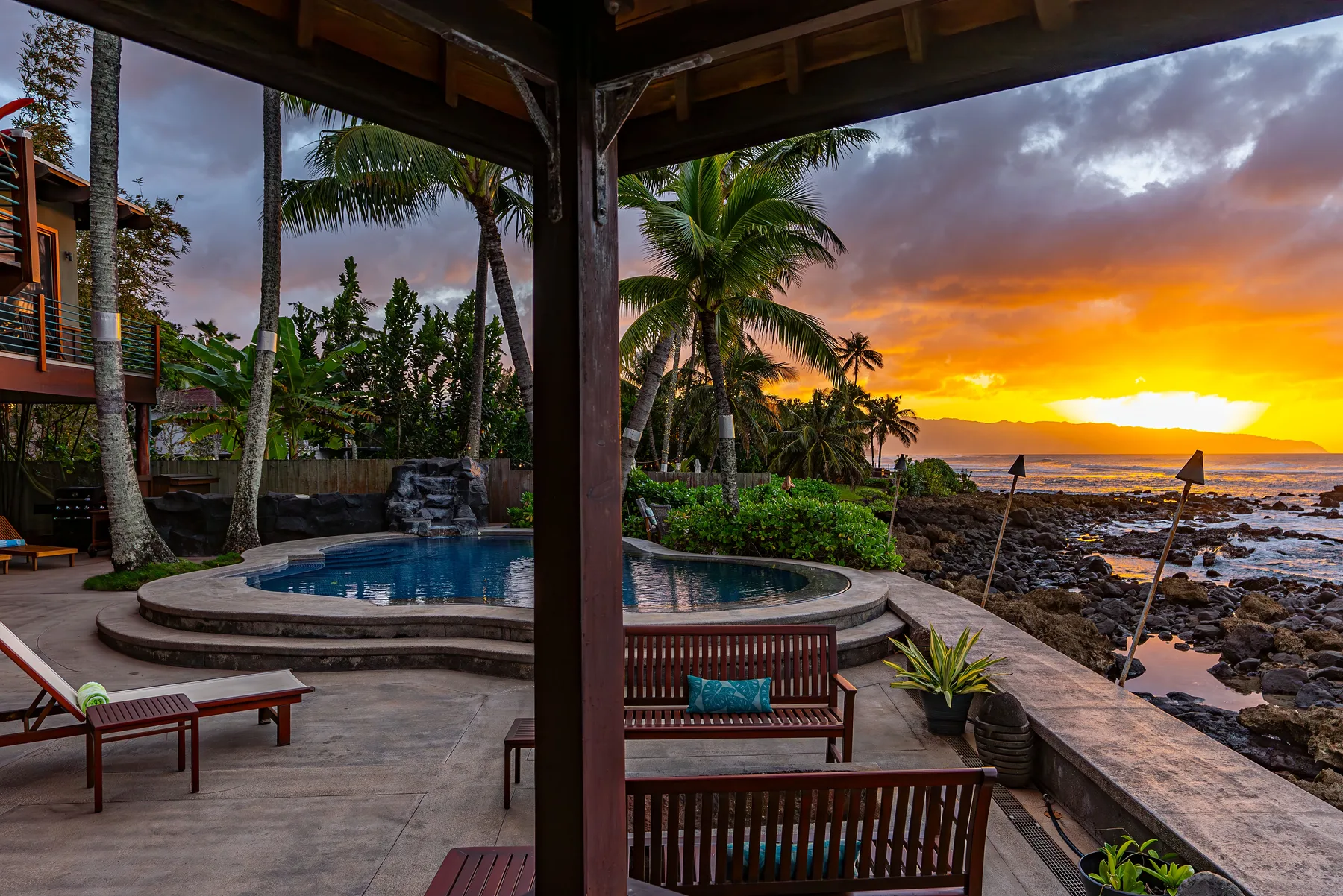 Un patio cubierto da a una piscina curva y una costa bordeada de palmeras al atardecer.