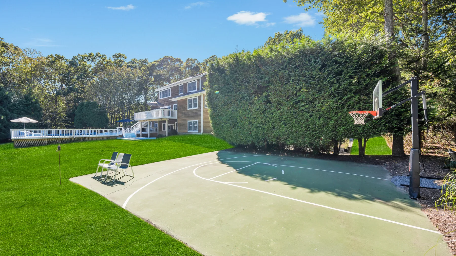 An outdoor basketball court borders a green lawn, with a hoop and chairs nearby.