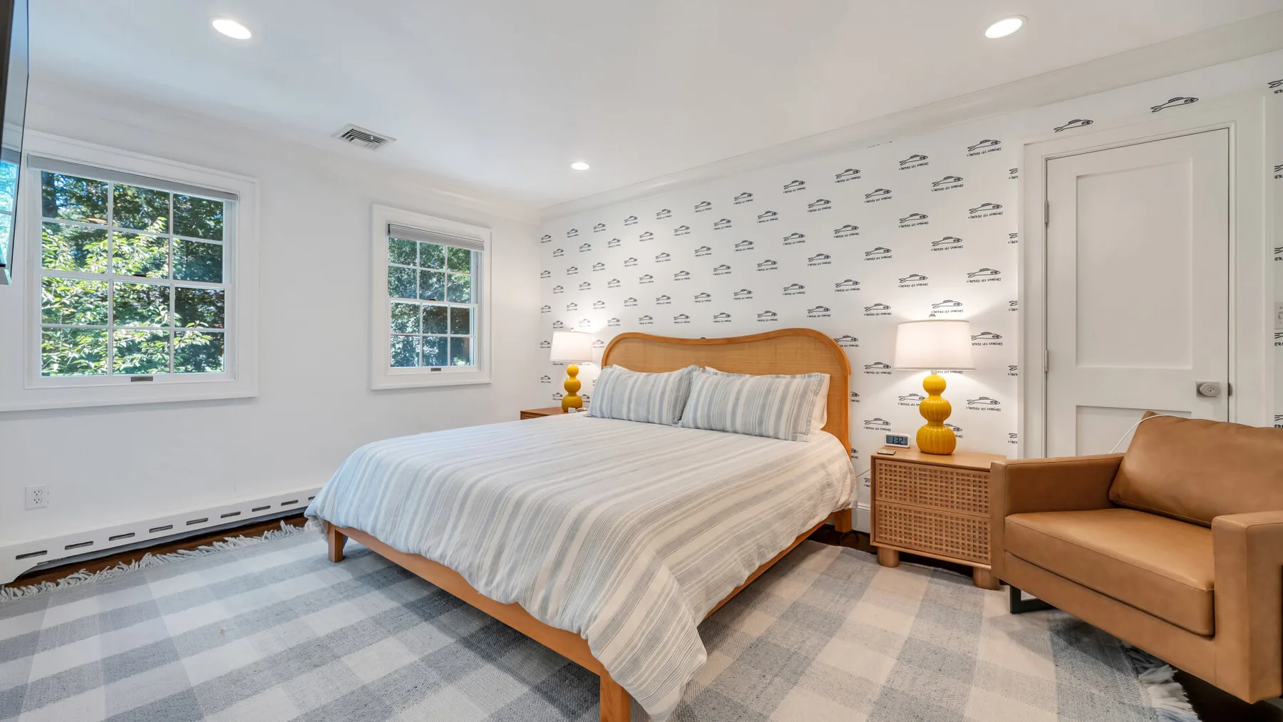 A bright bedroom with a bed, patterned wallpaper, and windows overlooking greenery.