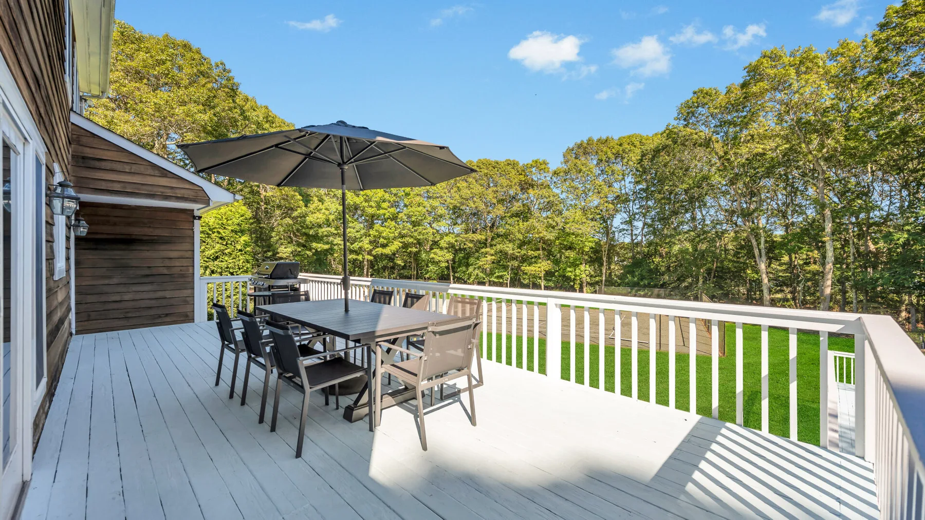 A wooden outdoor deck with a dining table and umbrella overlooks leafy trees.