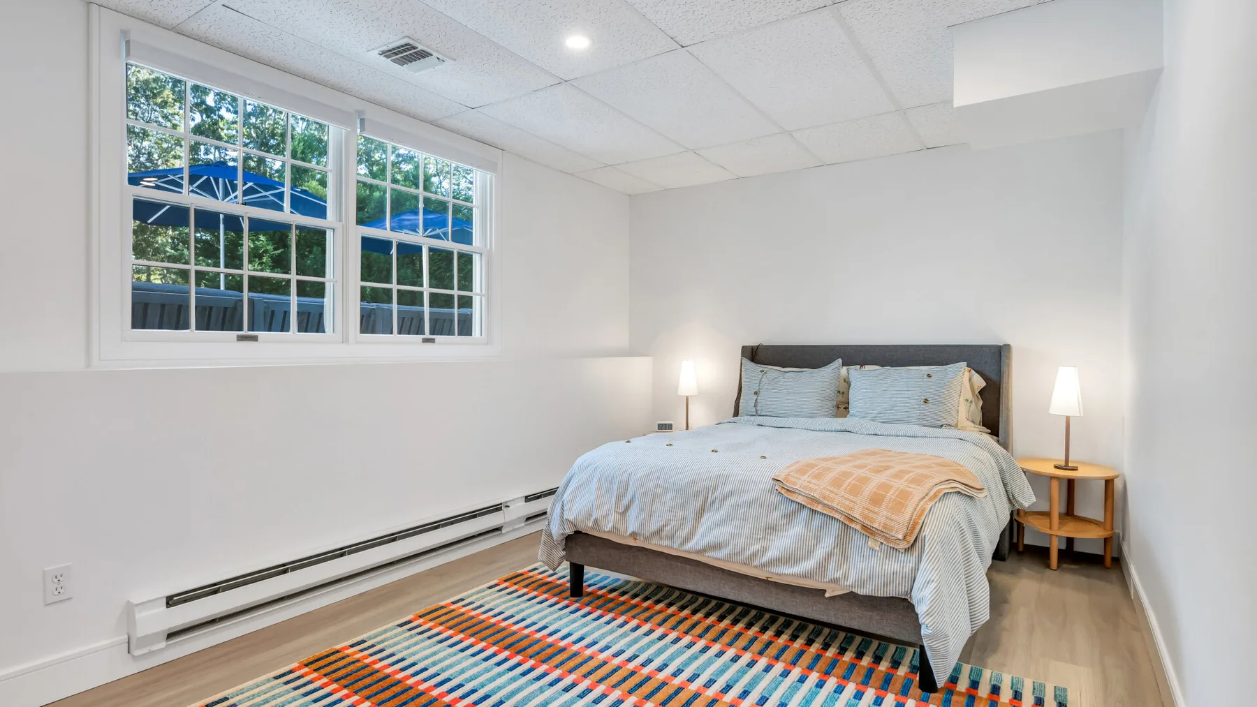 A bedroom with a bed beside large windows and a colorful striped rug.