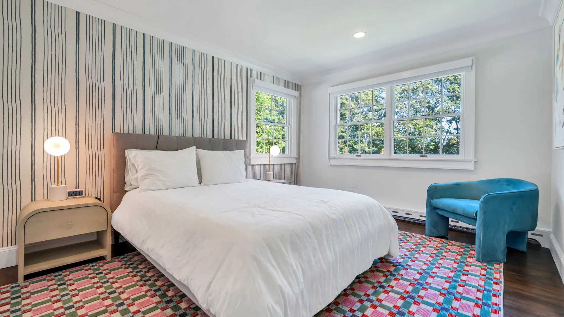 A bright bedroom with a large bed and blue seating beside the windows.