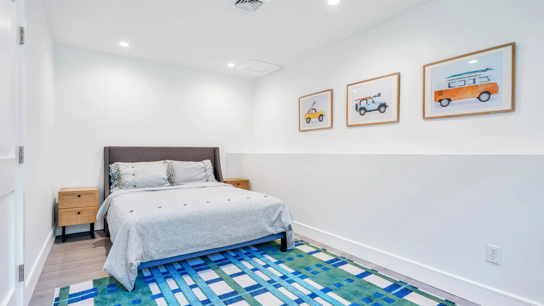 A bright bedroom with a bed, a blue patterned rug, and framed vehicle prints.