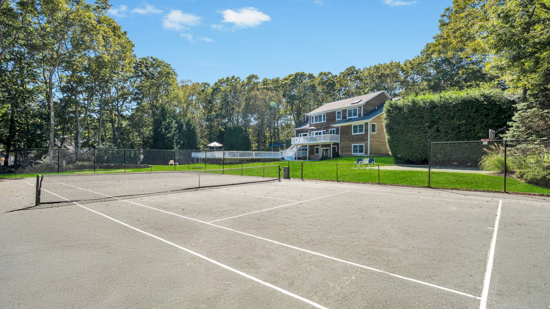 An outdoor tennis court sits beside a green lawn and a house.