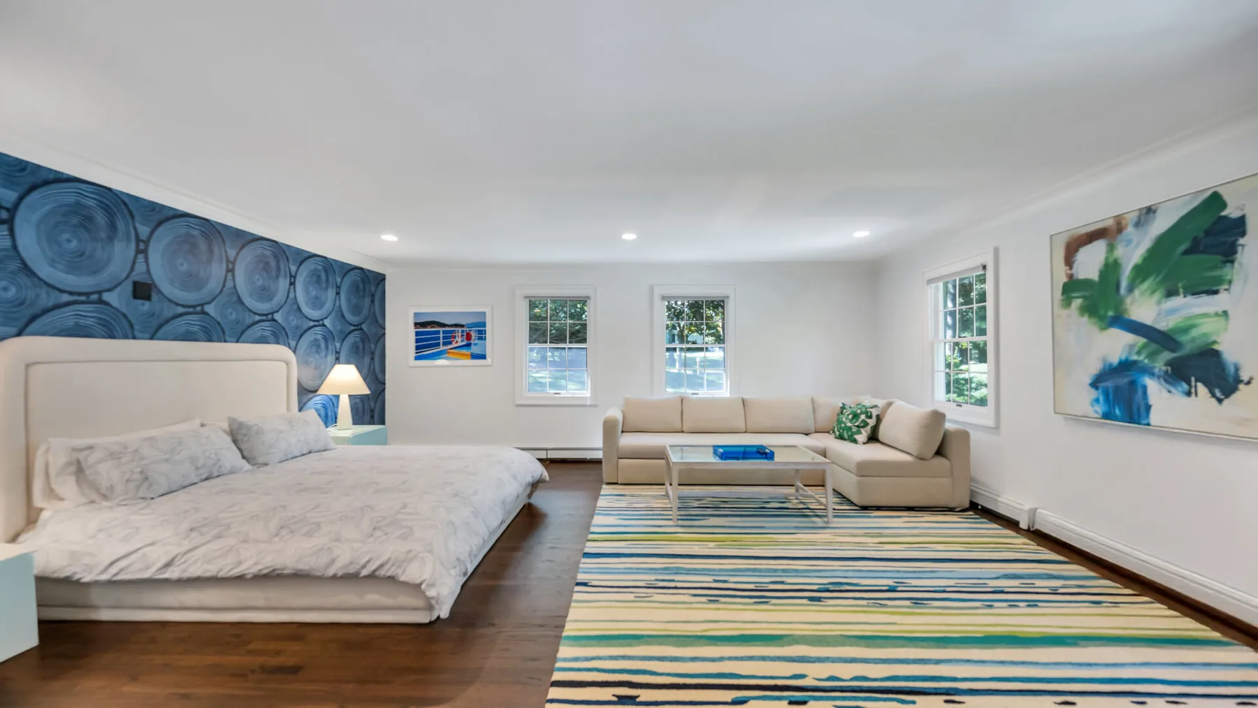 A bright bedroom and sitting area feature a bed, sectional seating, and a colorful striped rug.