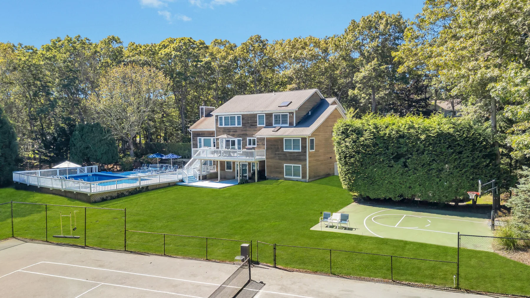 A house overlooks a broad green lawn with fenced sports courts and mature trees.