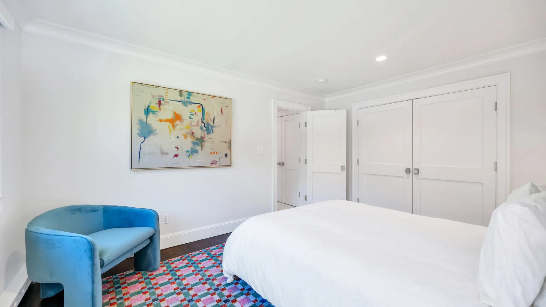 A bright bedroom with a white bed, blue seating, and colorful wall art.