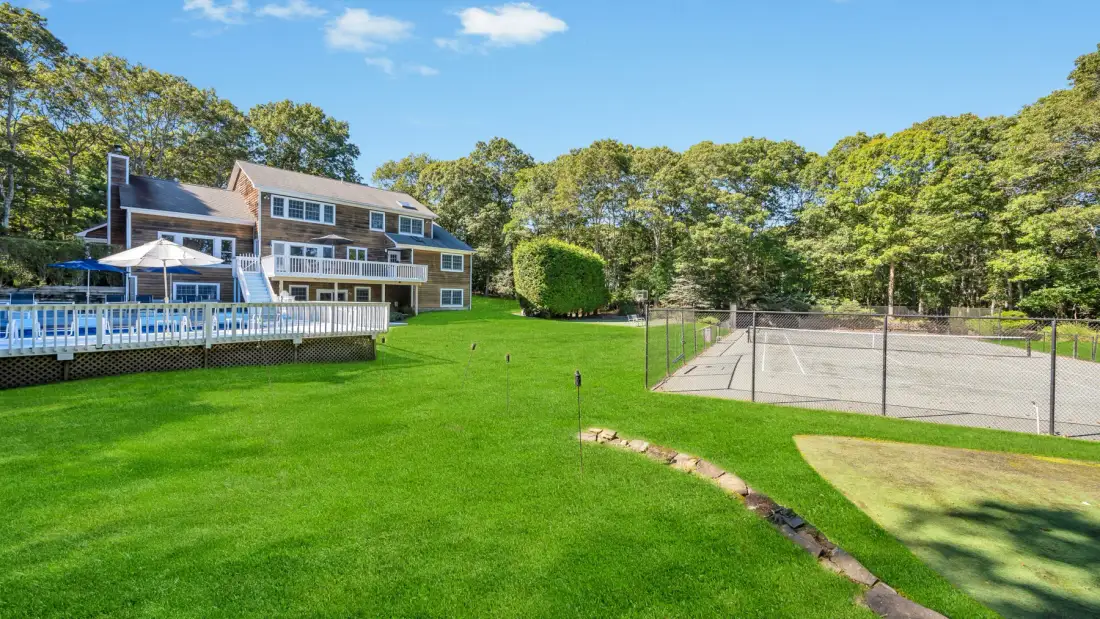 A large house overlooks a green lawn and a fenced tennis court.