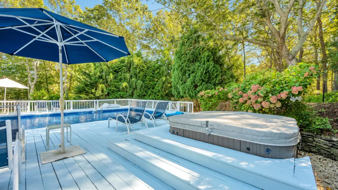 An outdoor pool area with a wooden deck, blue umbrella, and surrounding greenery.