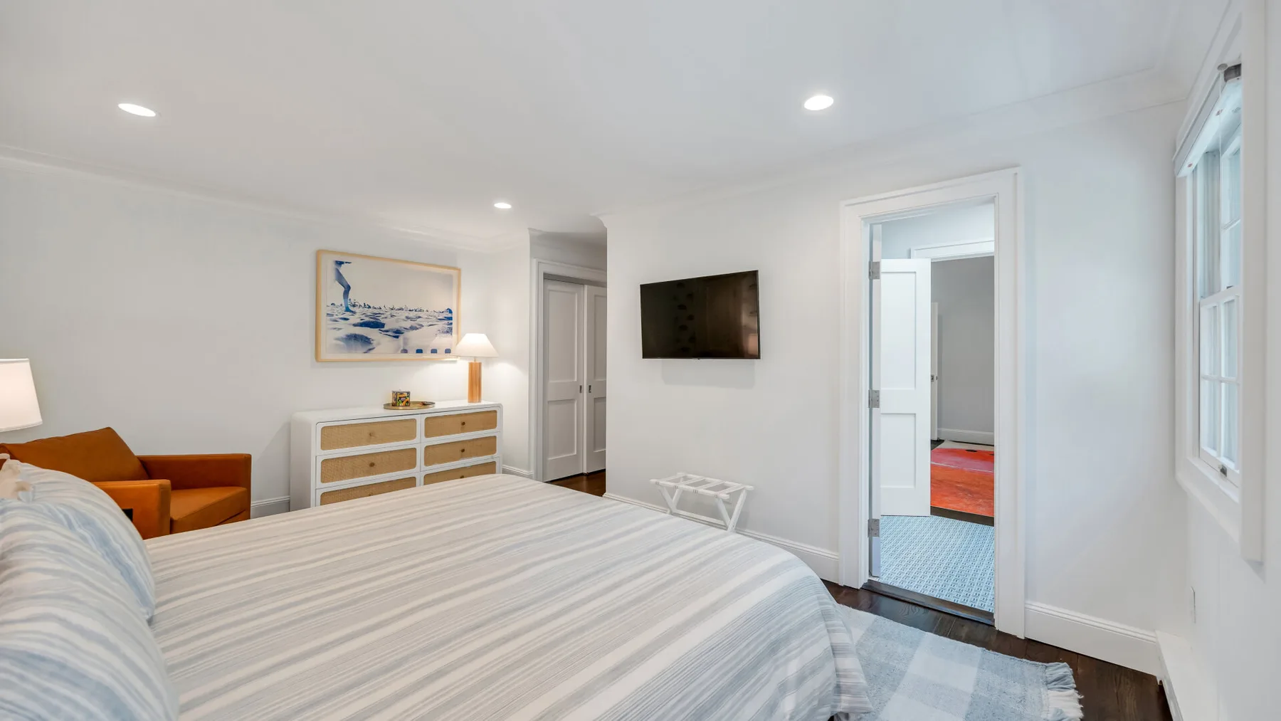 A bright bedroom with a striped bedspread, dresser, and wall-mounted television.