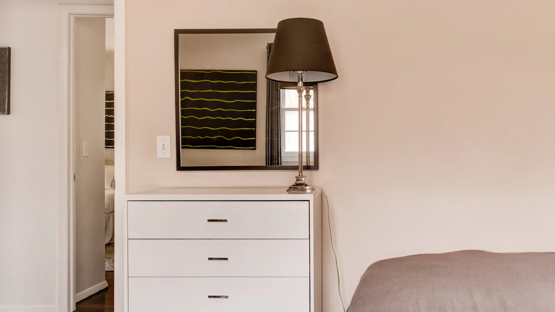 A bedroom corner with a white chest of drawers and a dark-shaded lamp.