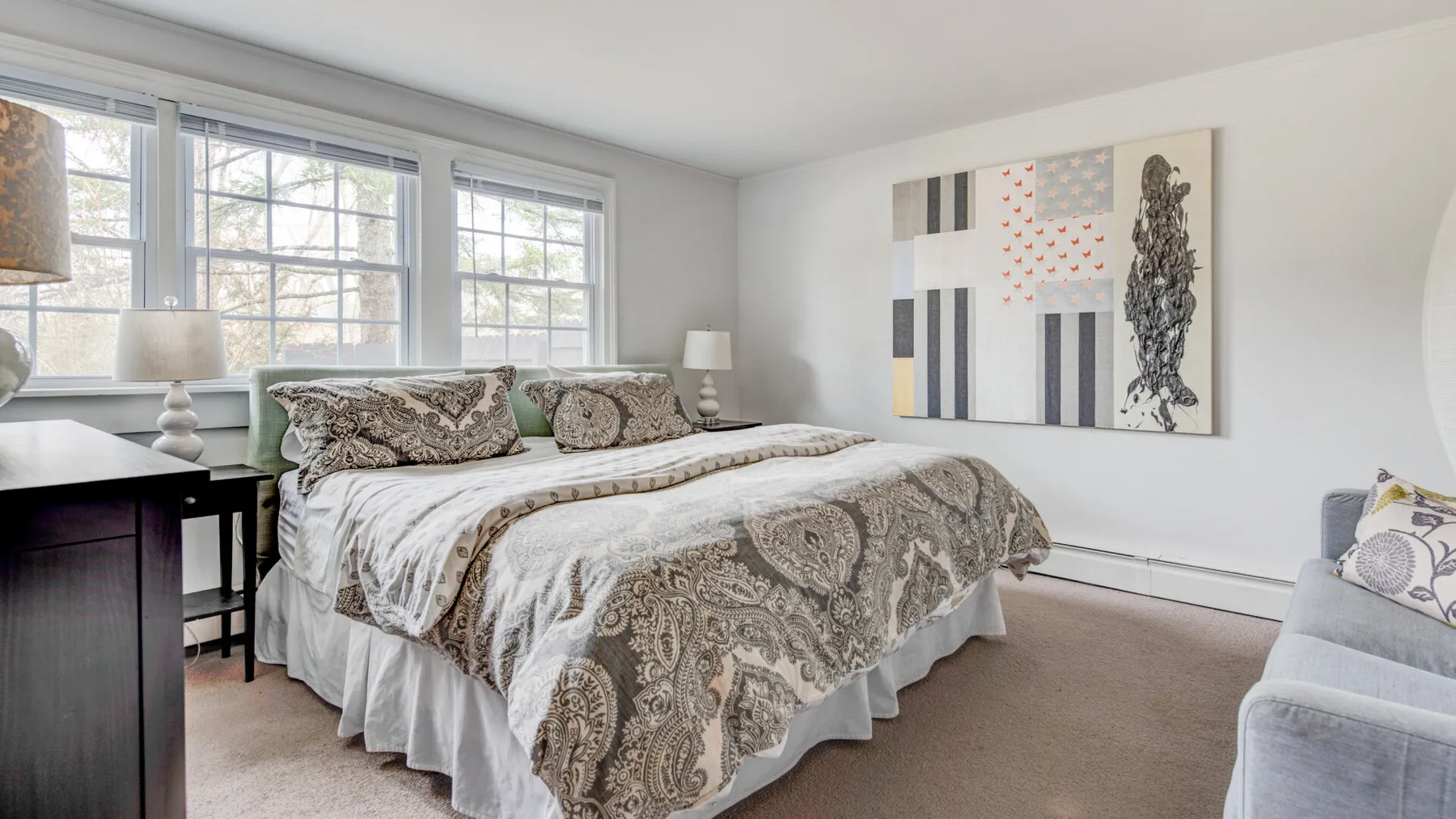 A bright bedroom with a patterned bedspread, large windows, and framed wall art.