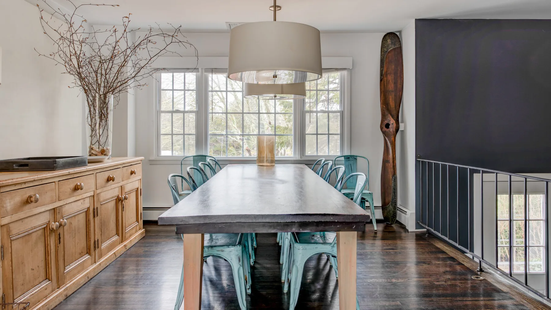 A dining area with a long table and blue-green seating in front of bright windows.