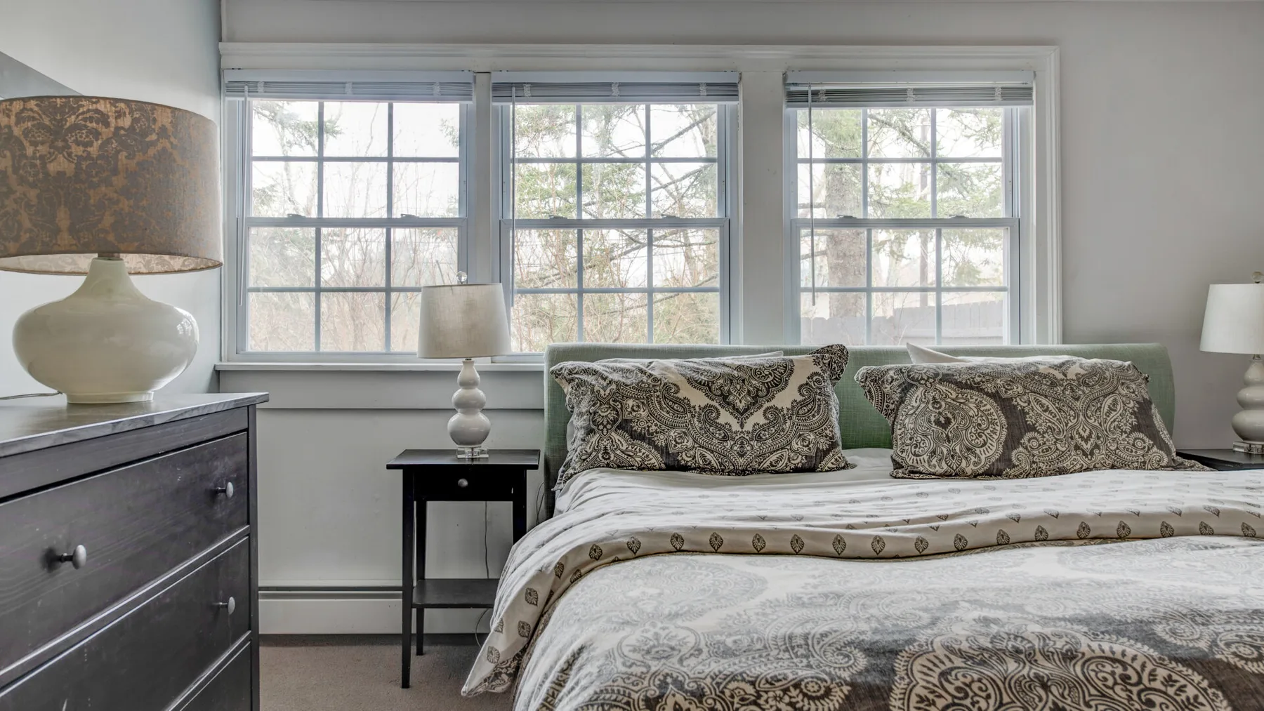 A bedroom with a neatly made bed and three large windows.