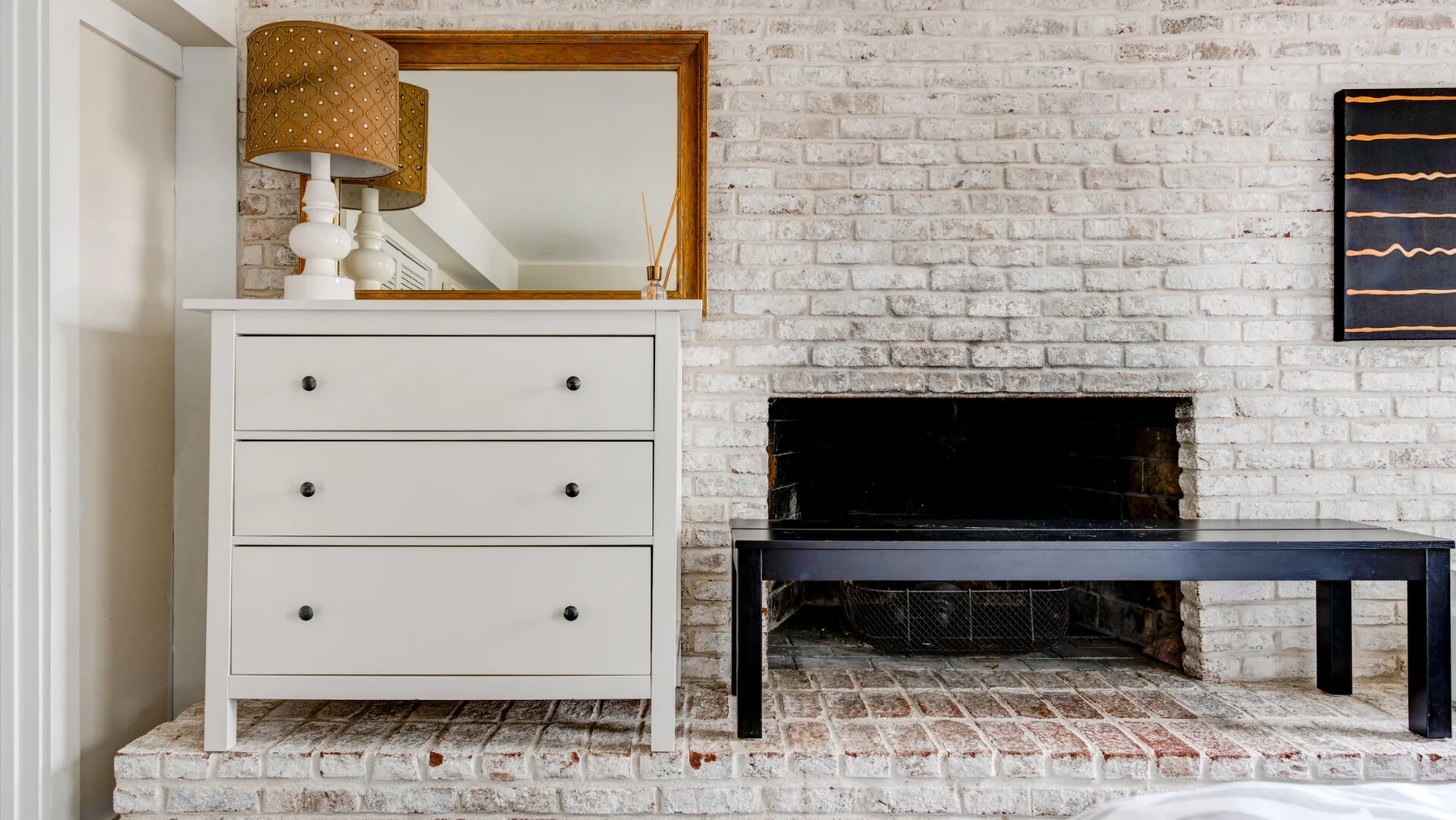 A living room with a white dresser, brick fireplace, and dark bench.