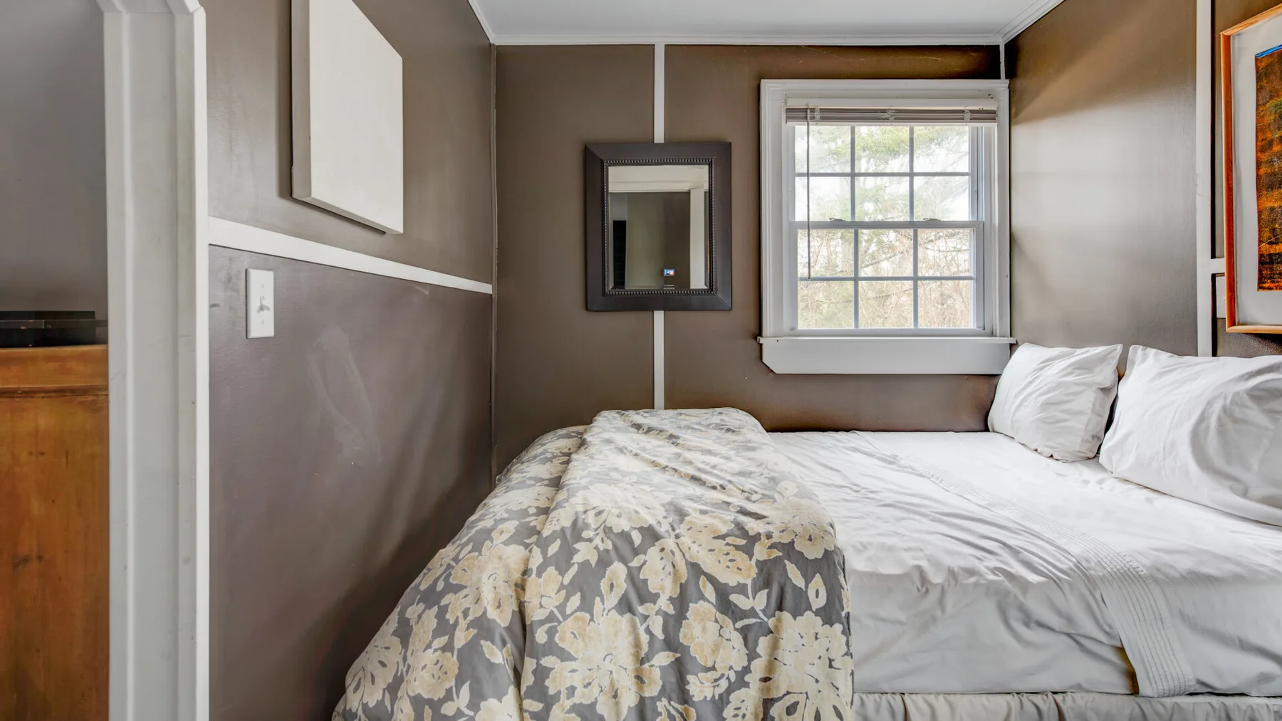 A bedroom with a bed beneath a window and a dark framed mirror.