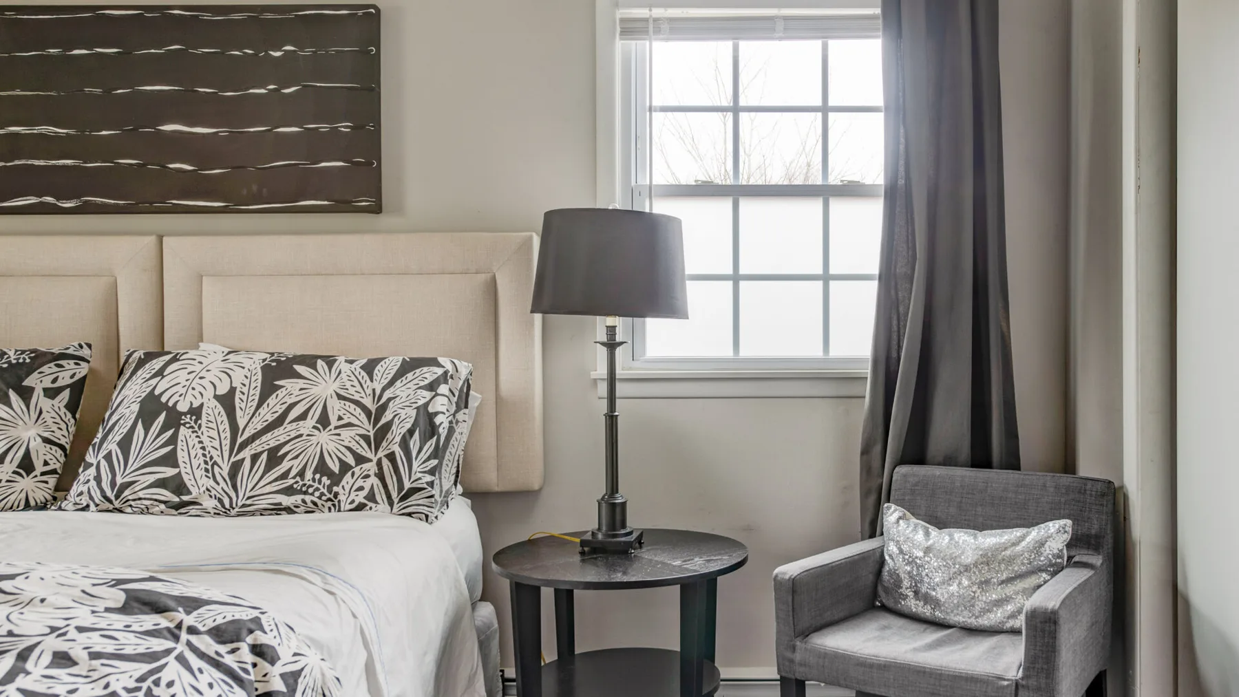 A bedroom with a bed, a bedside table, and a gray armchair beside the window.