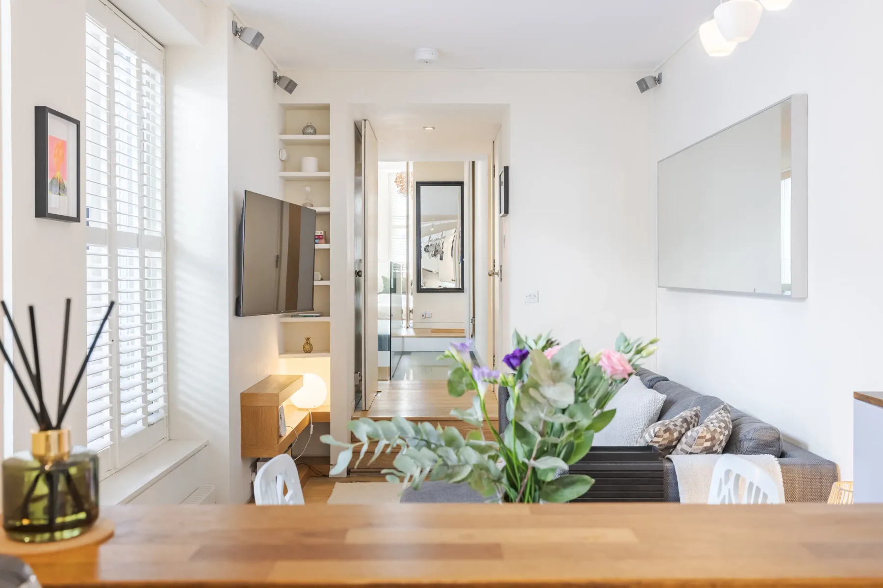 A bright living room with seating, a wall-mounted television, and a table in the foreground.
