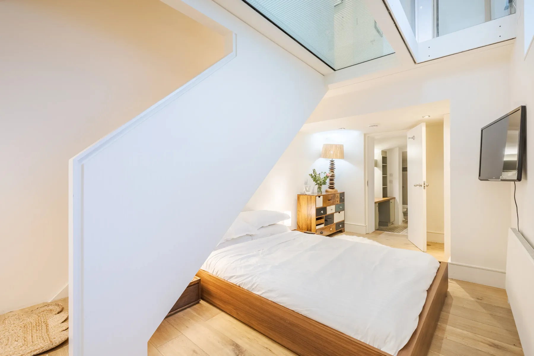 A bright bedroom with a bed beneath a staircase and a skylight overhead.