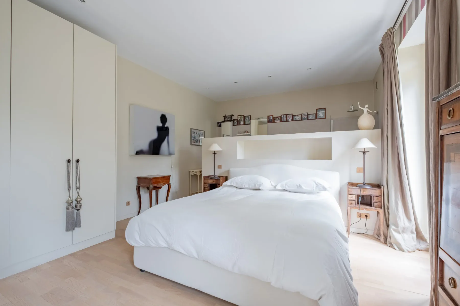 Un dormitorio luminoso con una cama blanca, mesas de noche y ventanas altas.