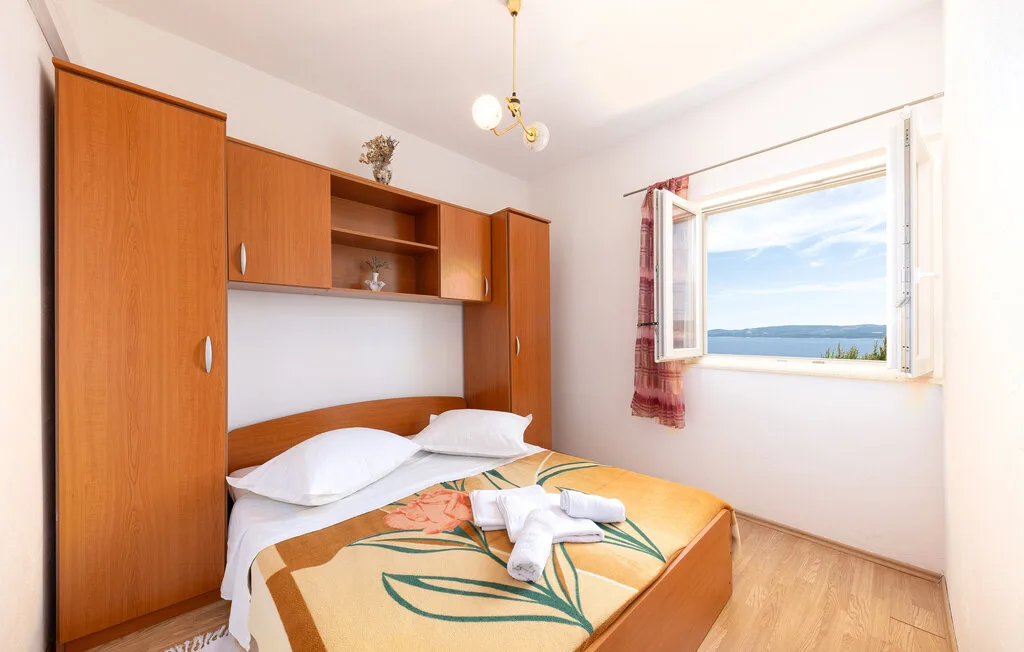A bedroom with a bed beneath wooden shelving and an open window overlooking the water.