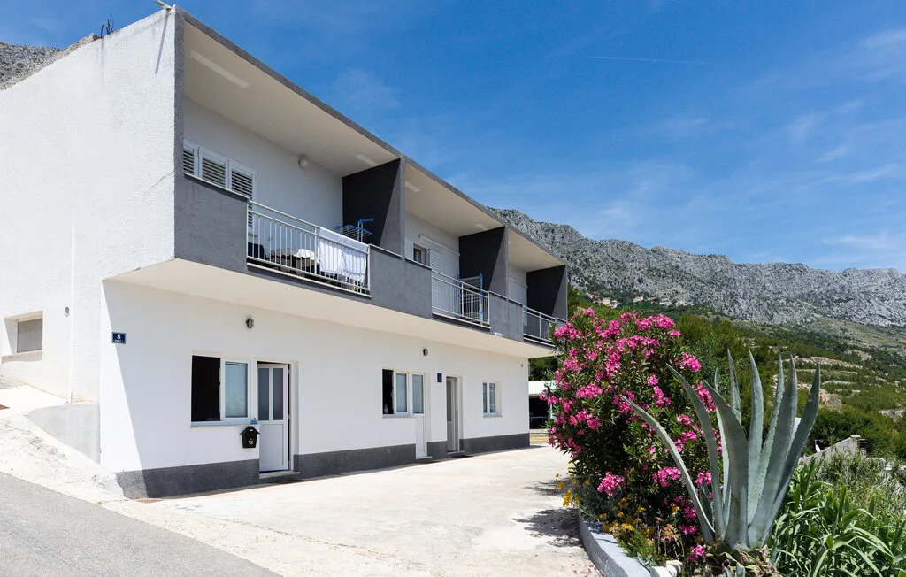 A white two-story building with balconies stands beside flowering plants against a mountain backdrop.
