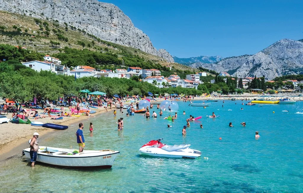 Una playa concurrida y zona de baño se encuentra bajo montañas rocosas y un pueblo en la ladera.