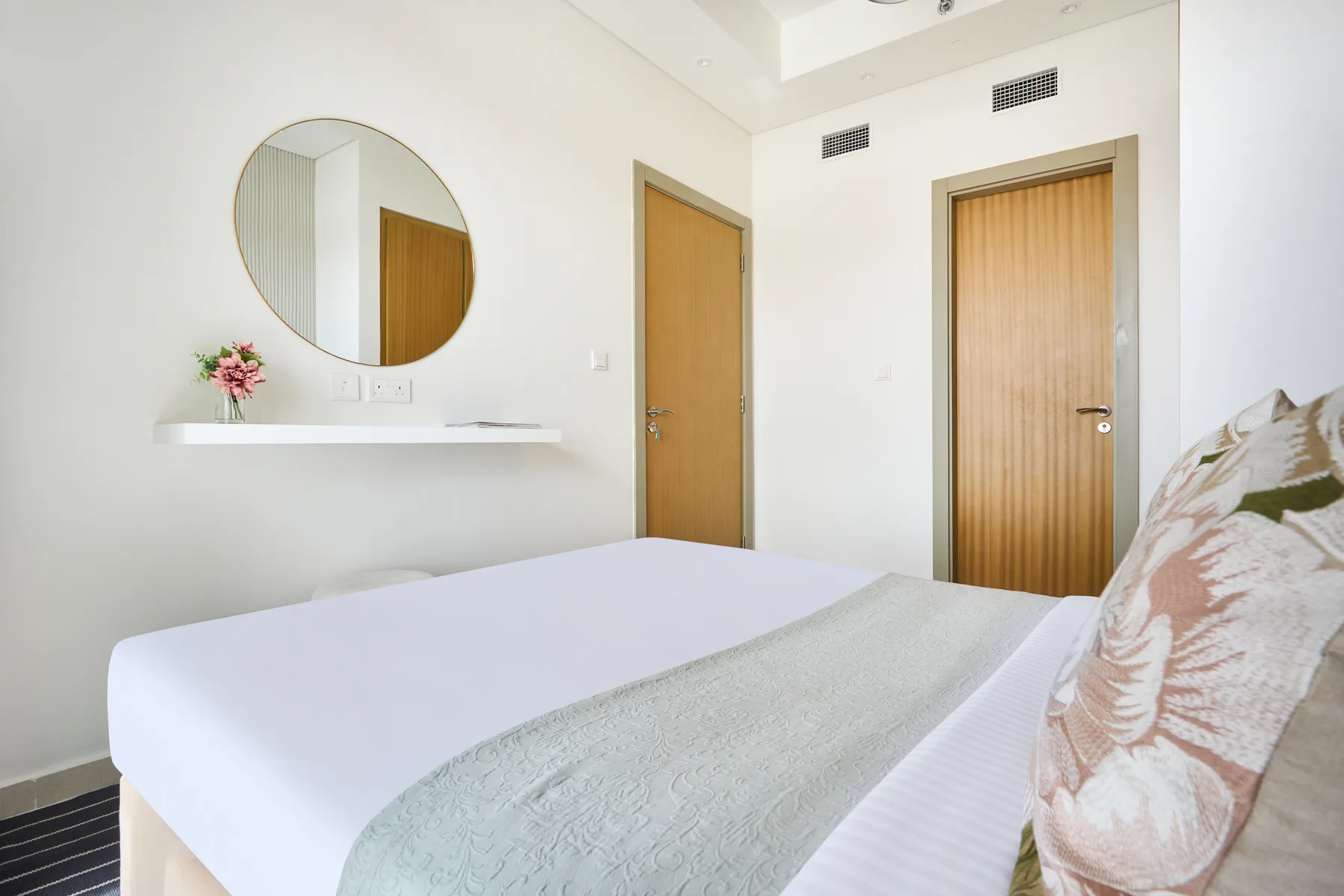 A bedroom with a neatly made bed and a round wall mirror.