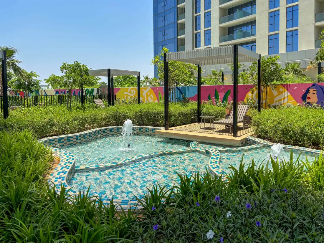 An outdoor pool with fountains sits beside a landscaped garden and modern building.