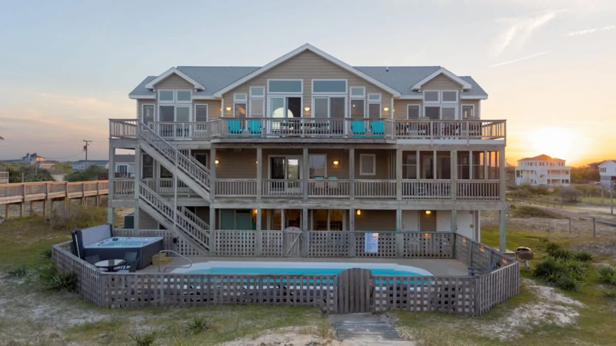 A large raised beach house with wraparound decks and an outdoor pool at sunset.