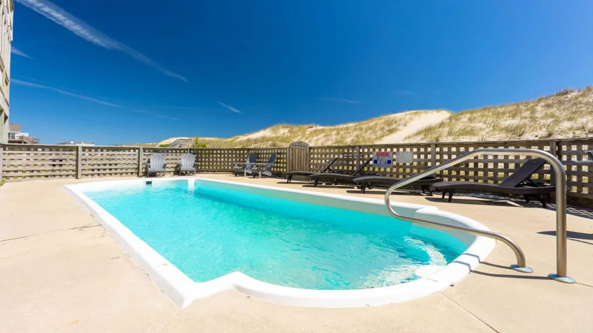 Una piscina al aire libre está junto a una cerca de madera y dunas de arena.