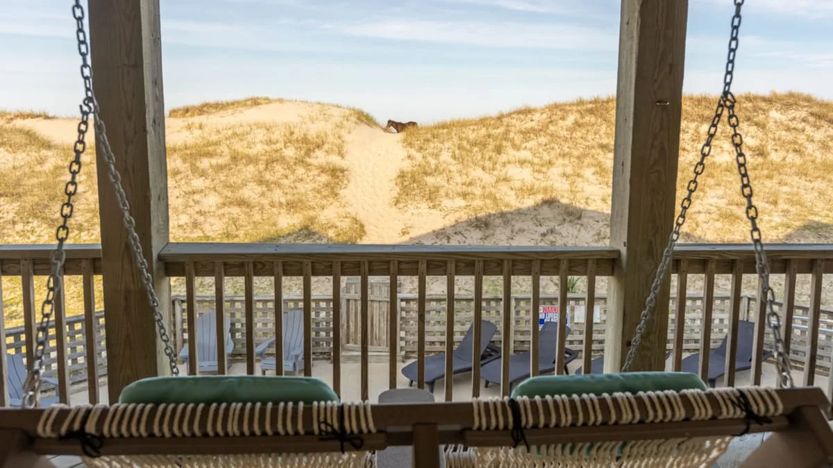 A wooden porch swing overlooks a sandy dune path and beachside buildings.