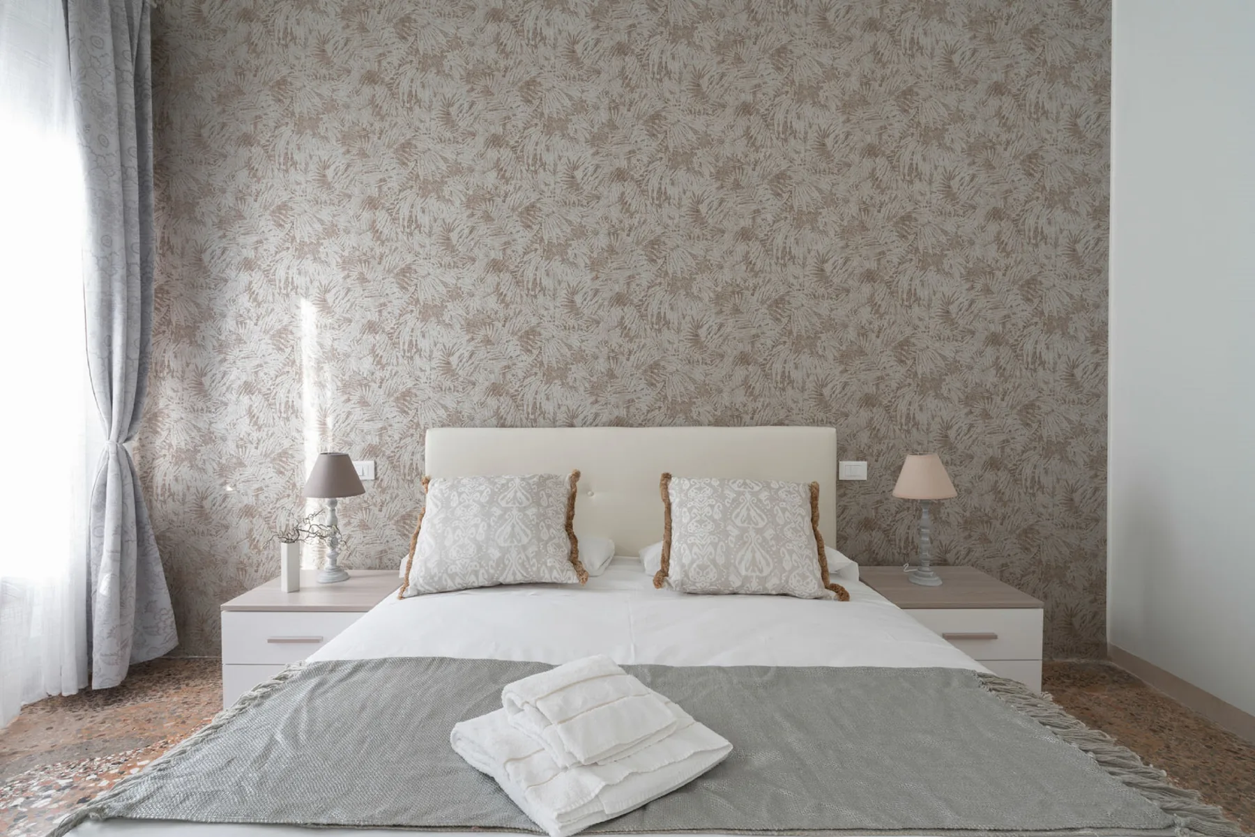 A bedroom with a neatly made bed, bedside lamps, and patterned wallpaper.