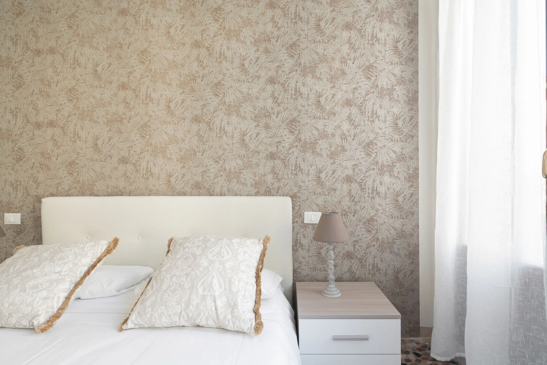 A bedroom with a bed, patterned pillows, and a bedside table beside a curtained window.