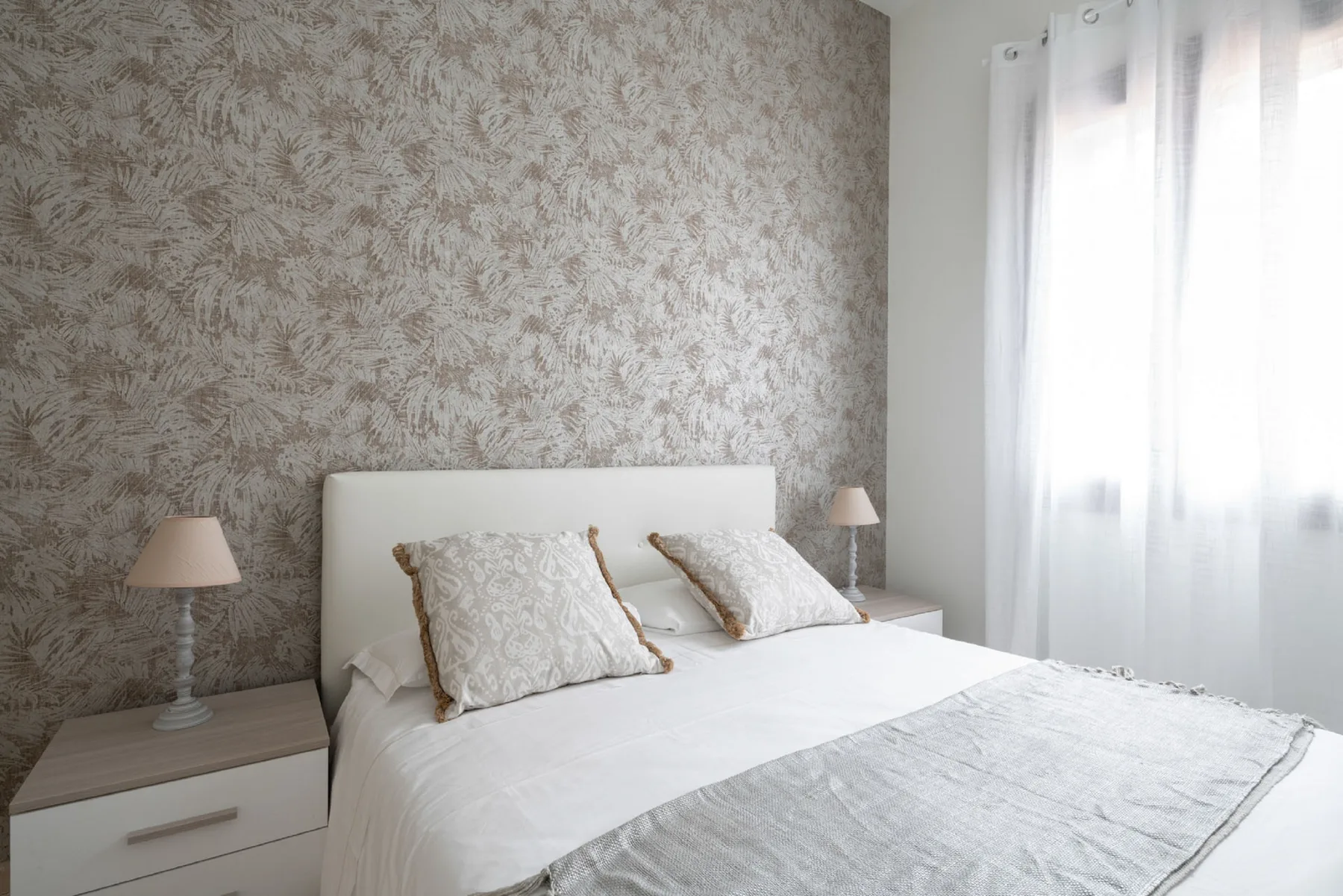 A bedroom with a white bed against a patterned wall and a bright curtained window.