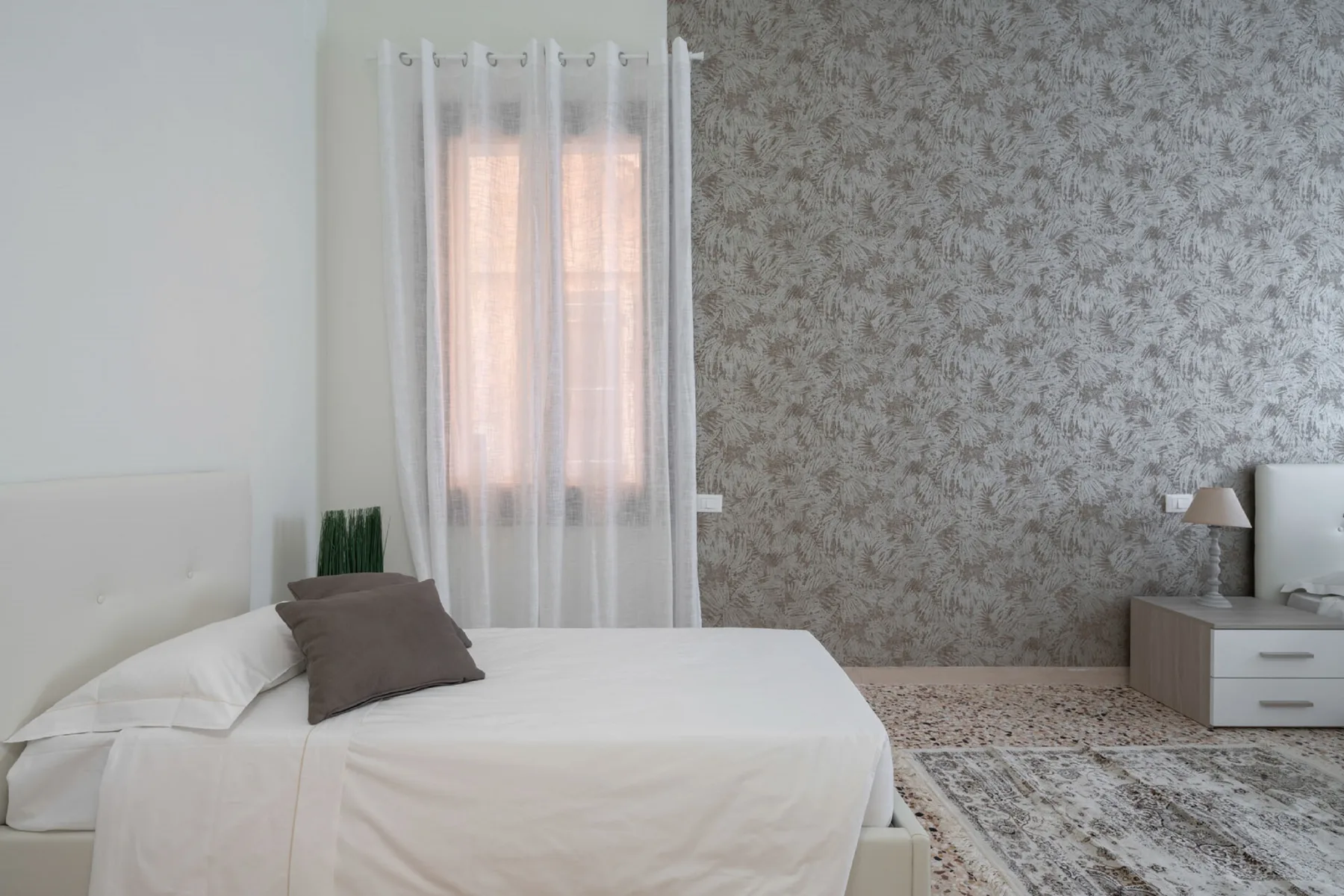 A bedroom with a bed beside a curtained window and patterned wallpaper.