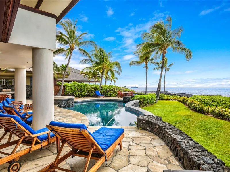 Un patio junto a una piscina tropical, con tumbonas azules, palmeras y vistas al mar.