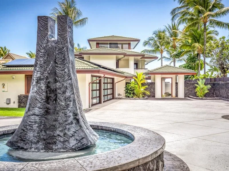 Una villa tropical se alza detrás de una fuente circular en un patio pavimentado.